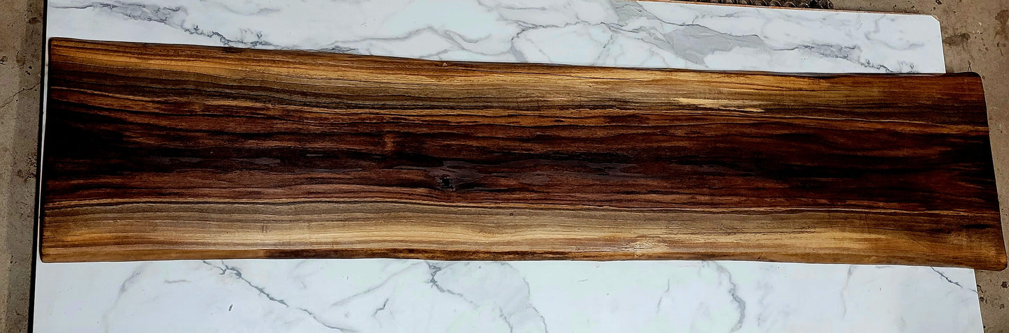 Extra long black walnut grazing board with light, medium, and dark streaks from the natural wood grain. Beautiful natural pattern creating an overall striking appearance with live wood edges. The grazing board is pictured on top of a granite table in the woodshop. 